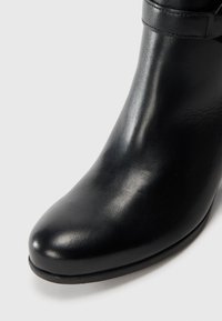 Bottine noire en cuir avec un design épuré, bout pointu et texture lisse. Elle dispose d'une simple sangle au niveau de la cheville pour un détail supplémentaire.