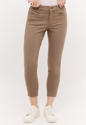 Slim-fit braune gekürzte Hose, getragen mit weißen Sneakers an einer Person, die vor einem einfarbigen Hintergrund steht.
