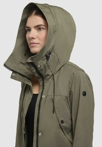 Veste à capuche vert olive avec un col montant, fermeture éclair et boutons-pression, cordons de serrage réglables et tissu texturé avec une finition mate.