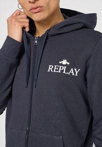 Marineblauwe rits-vest gemaakt van een katoenmix. Voorzien van een kenguruzak en een wit 'REPLAY' logo op de borst. Textuurafwerking.