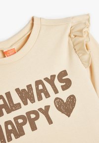 VAALEA BEIGE HUPARINE, JOSSA ON RAOBAKANKAAN TEKSTUURI, RÖYHEÄT HARTIAREUNAT JA TUMMA BRAUN "ALWAYS HAPPY" -TEKSTI SYDÄN KOROSTUKSELLA.