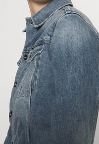 Chaqueta de mezclilla en azul claro con cierre de botones, cuello puntiagudo y dos bolsillos en el pecho. Presenta botones negros y un ajuste estructurado.