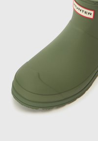 Stivale di gomma verde con punta arrotondata, suola texturizzata e un'etichetta rossa con la scritta "HUNTER" vicino alla parte superiore. Finitura liscia, altezza a metà polpaccio.