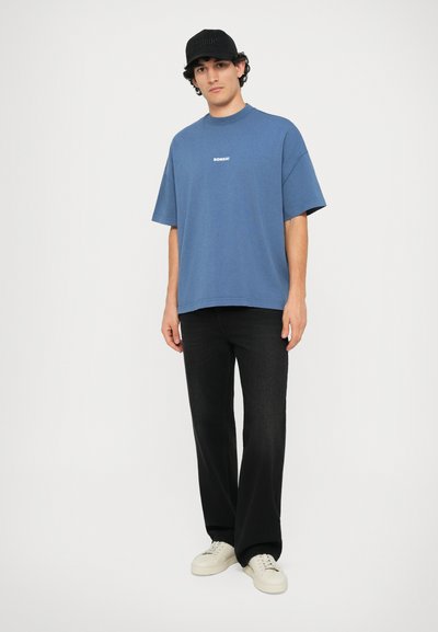 T-shirt bleu en coton avec un col rond et des manches courtes, présentant un logo "BONSÀI" blanc. Associé à un jean noir et des baskets blanches.