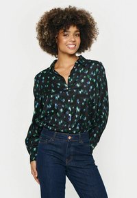 Camisa negra de botones con un patrón abstracto en verde y azul, de mangas largas y corte relajado, combinada con jeans azules oscuros.