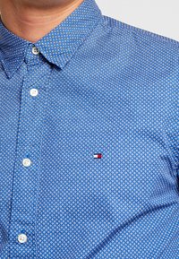 Blauwe button-up shirt met kleine witte geometrische patronen, voorzien van een gestructureerde kraag en een logopatch op de borst.