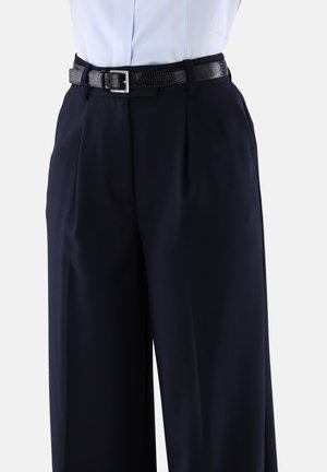 Person in einer hochtaillierten, weit geschnittenen navyblauen Hose mit vorderen Falten und einem schwarzen strukturierten Gürtel über einem in die Hose gesteckten weißen Hemd.