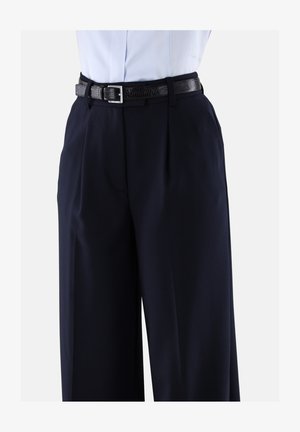 Person in einer hochtaillierten, weit geschnittenen navyblauen Hose mit vorderen Falten und einem schwarzen strukturierten Gürtel über einem in die Hose gesteckten weißen Hemd.