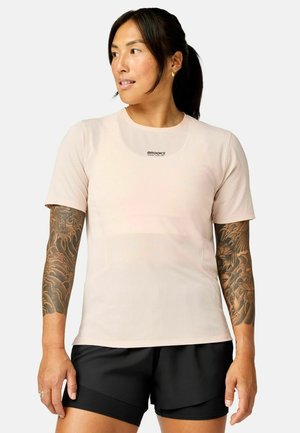 Kvinde med mørkt hår i en hestehale bærer en lysebeige kortærmet skjorte og sorte shorts, og viser tatoverede ærmer på begge arme.