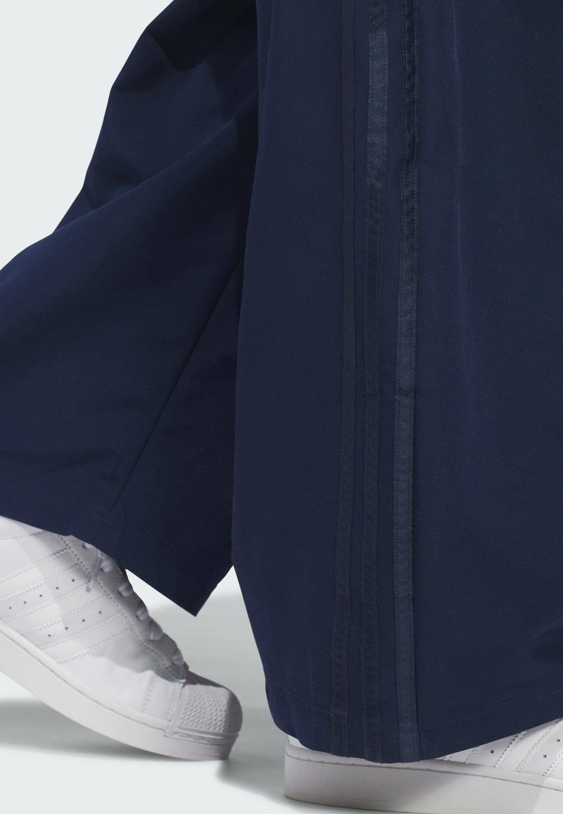 Zapatillas blancas combinadas con pantalones anchos de color azul marino con rayas verticales cosidas a lo largo de la costura exterior.