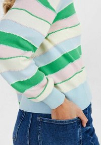 Person klädd i en pastellrandig tröja med gröna, rosa, blå och krämfärgade ränder, hand vilande i framlommen på ett par blå denimjeans.