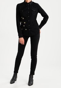 Camicia in denim nera con chiusura a bottoni, caratterizzata da decorazioni a forma di stella sul lato sinistro e due tasche sul petto. Abbinata a jeans skinny neri e stivali ankle.