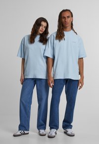 Camisetas oversized de color azul claro combinadas con jeans de mezclilla azules. Las camisetas tienen mangas cortas y un pequeño logo en el pecho. Calzado informal visible.