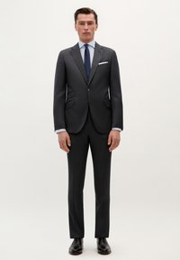 Traje gris oscuro con solapas en punta, dos bolsillos frontales y un corte ajustado. Combinado con una camisa azul clara y una corbata azul marino. Zapatos de vestir negros.