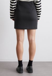Zwarte stoffen mini-jurk met achterste plooien, gecombineerd met zwarte sokken en donkere schoenen. Model draagt eengestreepte lange mouw shirt.