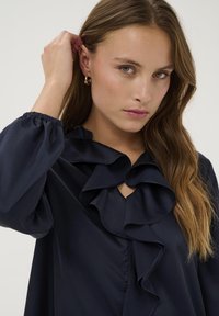 Donkerblauwe blouse met een gerimpelde kraag en een sleutelgatdetail, gemaakt van gladde stof met elastische manchetten. Het model heeft golvend haar en gouden oorbellen.