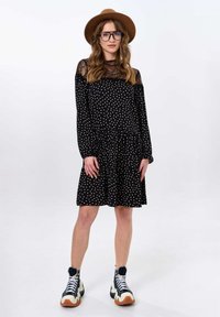 Zwarte jurk met lange mouwen en kanten halslijn, met witte stippen. Gecombineerd met een bruine hoed en chunky zwart-witte sneakers.