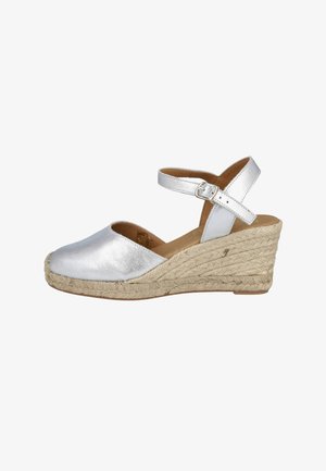 Sandalia cuña de cuero plateado con correa de tobillo con hebilla y suela de yute trenzado sobre fondo blanco.