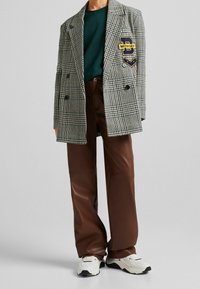 Blazer gris de cuadros de perro de presa de doble botonadura con un parche amarillo de "Día de Selección", combinado con pantalones marrones de piel sintética y zapatillas blancas.