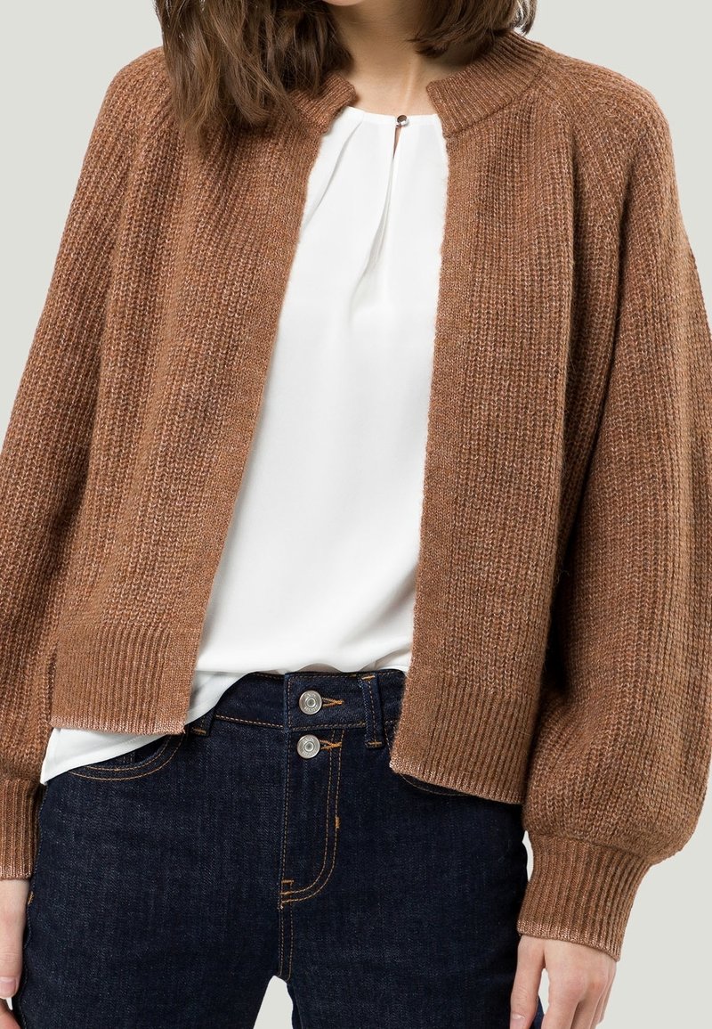 Femme portant un cardigan marron en tricot sur un chemisier blanc et un jean bleu foncé taille haute avec une fermeture double bouton.