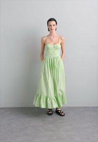 Groene geruite maxi-jurk met een aansluitend lijfje en een gerimpelde zoom, ondersteund door dunne banden. Draag het met zwarte sandaaltjes op een lichtgrijze vloer.