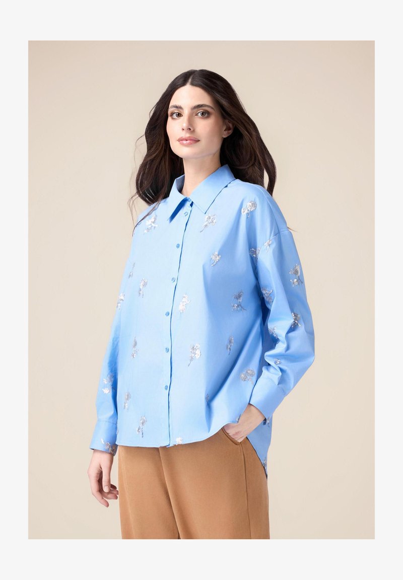 Light blue button-up shirt made of smooth fabric, featuring silver floral embroidery and a relaxed fit with long sleeves and a pointed collar.