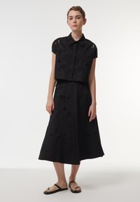 Black two-piece outfit consisting of a cropped, embroidered short-sleeve top and a flowing, mid-length skirt with similar detailing. Sandals are worn.