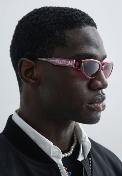 Κόκκινα διαφανή γυαλιά ηλίου με σκοτεινούς φακούς, γωνιώδη σχήματα και χαραγμένες λεπτομέρειες στον βραχίονα. Διαθέτουν ξεχωριστό σχεδιαστικό στοιχείο αντίθεσης.