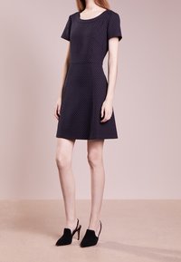 Vestido ajustado de manga corta en color azul marino con una tela texturizada que presenta pequeños puntos blancos. La falda se ensancha desde la cintura. Usado con tacones negros.