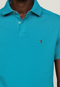 Turkusowa koszulka polo wykonana z teksturowanej tkaniny. Posiada kołnierz, dwa guziki i małą markową emblemat w kolorach czerwonym, białym i niebieskim.