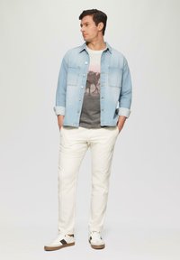 Veste en denim bleu clair sur un T-shirt graphique gris. Pantalons blancs et sneakers décontractés avec un accent marron. Modèle se tenant avec les mains dans les poches.