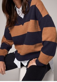 Pull en V rayé alternant bleu marine et beige, texture tricotée et poignets côtelés. Porté par-dessus une chemise blanche à col.