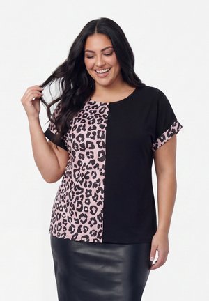 Donna che sorride con capelli lunghi e scuri indossa una maglietta a maniche corte, metà nera tinta unita e metà rosa con stampa leopardata, abbinata a una gonna in pelle nera.