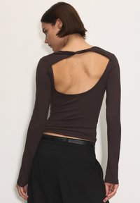 Brązowa bluzka z długim rękawem i otwartymi plecami, z detalem skrętu przy dekolcie. Gładki, dopasowany materiał kontrastuje z czarnymi spodniami.