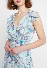 Femme portant une robe sans manches bleu clair avec un imprimé floral violet et sarcelle, agrémentée de détails froncés le long du col et de la taille.