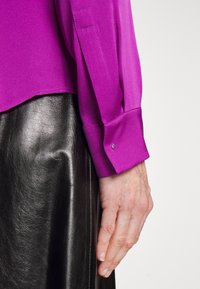 Handen vilar vid sidan, klädd i en lila långärmad skjorta med manschettknappar och en svart strukturerad läderkjol.