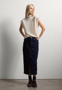 Top sin mangas de un beige claro con un cuello alto, combinado con una falda midi de mezclilla oscura que presenta una abertura frontal y detalles en bronce. Zapatos marrones añaden contraste.