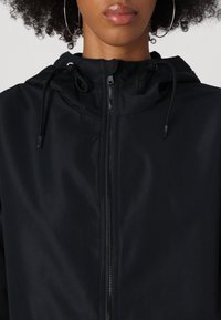 Černá mikina s kapucí hladkého vzhledu, vybavená předním zipem, šňůrkami na krku a minimalistickým designem.