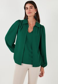 Blusa verde de manga larga con cuello tipo camisa y mangas voluminosas, que presenta una textura suave y una silueta estructurada.
