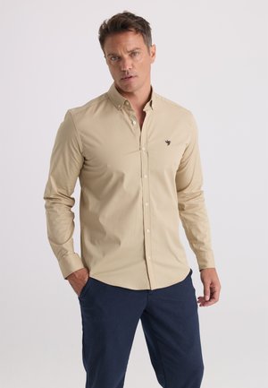 Homme portant une chemise beige boutonnée avec un petit logo d'oiseau et un pantalon bleu foncé sur un fond clair uni.