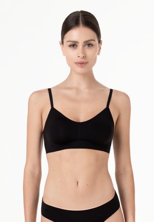 Giovane donna con i capelli raccolti che indossa un reggiseno nero senza cuciture e biancheria intima coordinata su uno sfondo bianco uniforme.