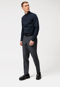 Chemise à manches longues marine, boutonnée, assortie à un pantalon à carreaux gris. Le mannequin se tient debout avec des chaussures de ville noires et une ceinture noire. Texture de tissu lisse.