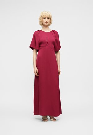 Kvinne med kort, krøllete blondt hår som har på seg en magenta kjole som går helt ned til gulvet med korte puffede ermer og åpne hæler, stående foran en ensfarget bakgrunn.