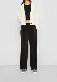 Krämfärgad ribbad cardigan med knappar fram, svart kort topp, svarta veckade vida byxor och skor med rutigt mönster.