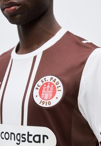 Κοντινό πλάνο ενός ατόμου που φοράει τη φανέλα της FC St. Pauli με καφέ και λευκές ρίγες, το έμβλημα του συλλόγου και το λογότυπο χορηγού ορατά στο στήθος.