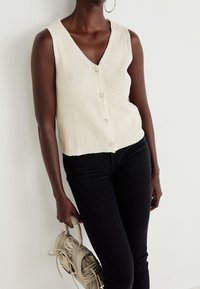 Krämfärgad stickad väst med V-hals, med knäppning fram och ribbat mönster. Matchas med svarta tajta jeans och en handväska.