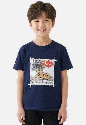 Garçon souriant portant un t-shirt bleu marine avec le texte graphique "La vie est meilleure à la plage" et une flèche étiquetée "OCÉAN" sur un fond blanc.