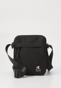 Sac bandoulière noir en tissu durable, doté d'une fermeture zippée en haut, d'une bandoulière ajustable et d'un petit patch logo Champion sur le devant.