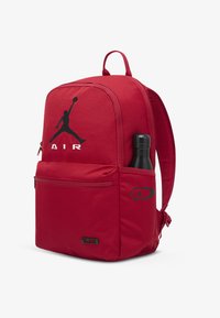 Κόκκινο σακίδιο με λείο ύφασμα, με μαύρο λογότυπο Jordan και  κείμενο «AIR». Η πλαϊνή τσέπη κρατάει μαύρο παγούρι νερού. Ρυθμιζόμενοι ιμάντες ώμου.