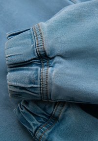 Tela de mezclilla azul clara con puños fruncidos. Costura notable en un color marrón claro que contrasta a lo largo de la costura. Textura suave, diseño casual.